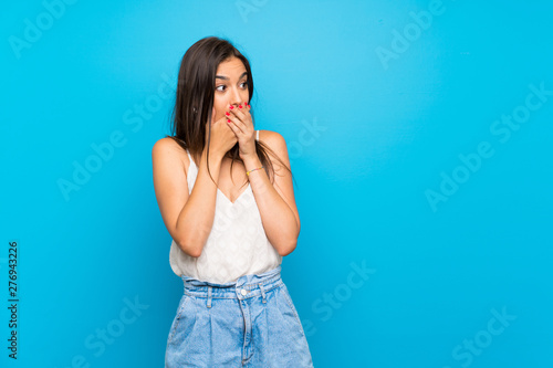 Young woman over isolated blue background covering mouth and looking to the side