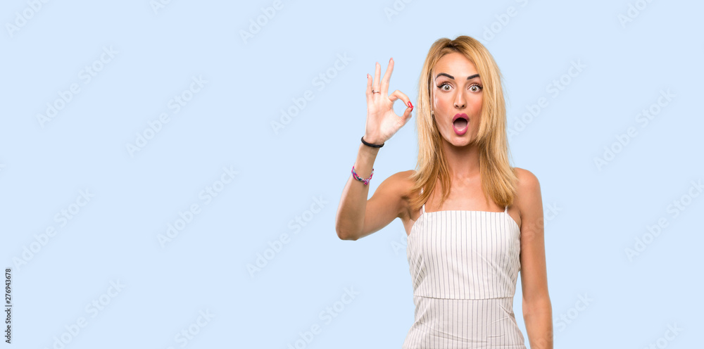 Young blonde woman surprised and showing ok sign over isolated blue background