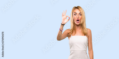 Young blonde woman surprised and showing ok sign over isolated blue background