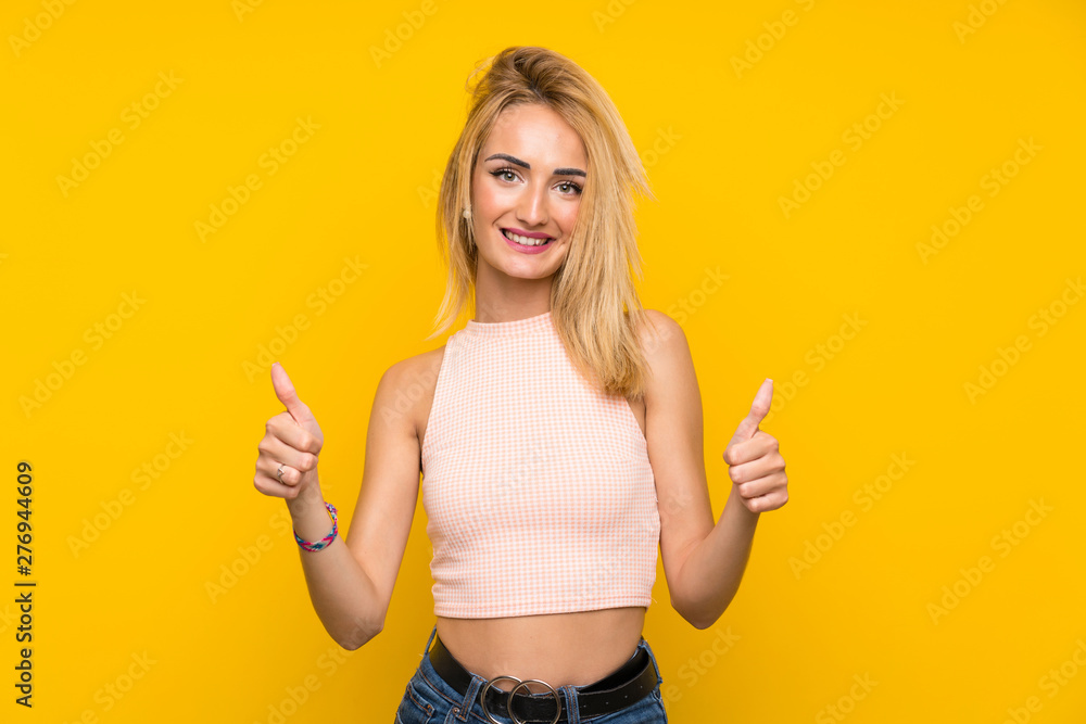 Young blonde woman over isolated yellow wall giving a thumbs up gesture Young blonde woman over isolated yellow wall giving a thumbs up gesture