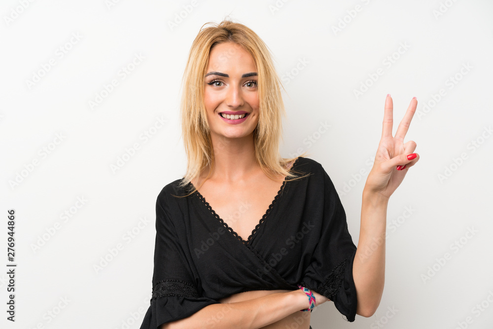 Young blonde woman over isolated white wall smiling and showing victory sign Young blonde woman over isolated white wall smiling and showing victory sign
