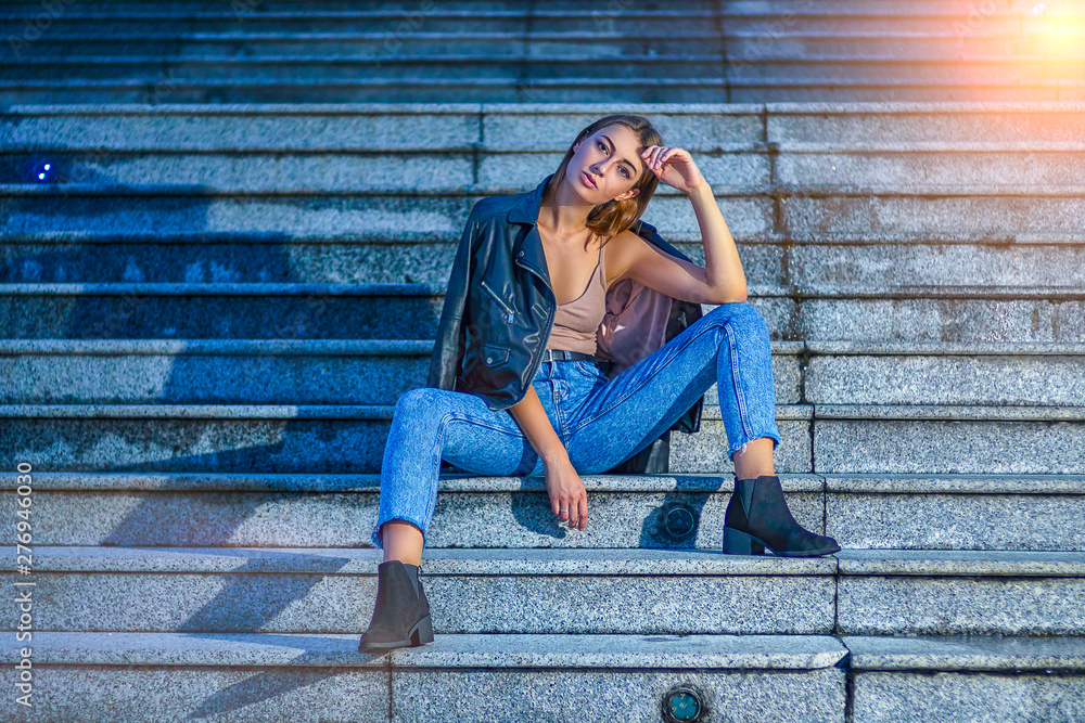 Sexy young female model is fashionable posing sitting on steps in denim and leather jacket. Fashion street photo shoot with beautiful girl Sexy young female model is fashionable posing sitting on steps in denim and leather jacket. Fashion street photo shoot with beautiful girl