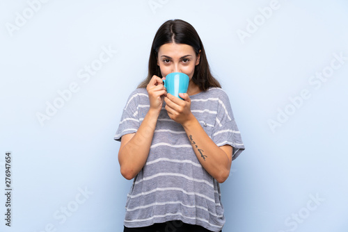 Young brunette woman over isolated blue background holding hot cup of coffee
