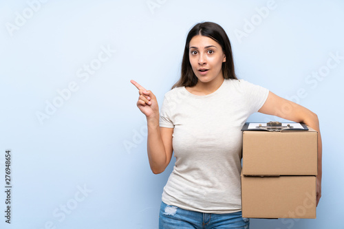 Young delivery woman over blue brick wall surprised and pointing side
