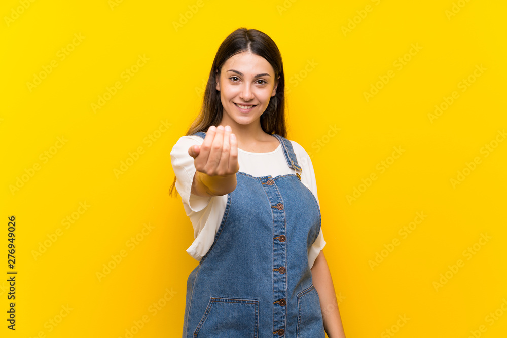 Young woman in dungarees over isolated yellow background inviting to come with hand. Happy that you came Young woman in dungarees over isolated yellow background inviting to come with hand. Happy that you came