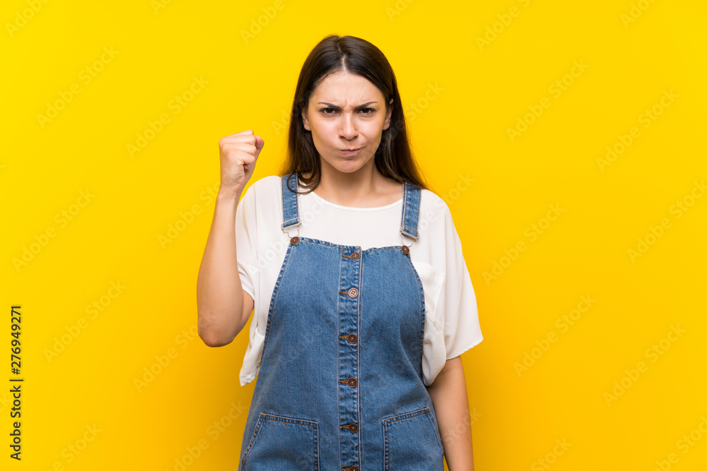 Young woman in dungarees over isolated yellow background with angry gesture Young woman in dungarees over isolated yellow background with angry gesture