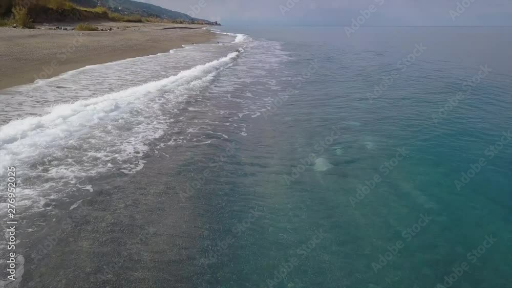Drone flies over sea coast, aerial view of waves reaching shore and splashing breaking on empty pebble beach, mountains on horizon, pan camera movement Drone flies over sea coast, aerial view of waves reaching shore and splashing breaking on empty pebble beach, mountains on horizon, pan camera movement