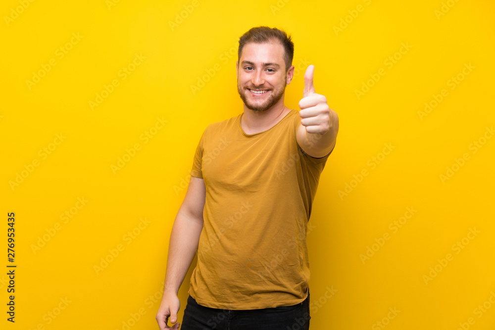 Blonde man over isolated yellow wall with thumbs up because something good has happened Blonde man over isolated yellow wall with thumbs up because something good has happened