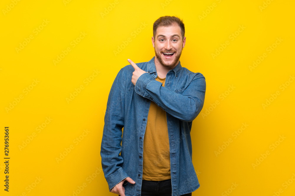 Blonde man over isolated yellow wall surprised and pointing side Blonde man over isolated yellow wall surprised and pointing side
