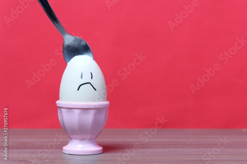 A boiled egg with a chopped shell stands in a poached. On the egg painted smiley. A spoon is stuck in it for food. Against the background of coral color.