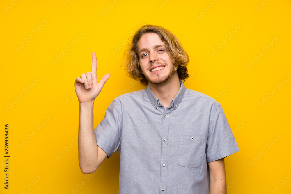 Blonde man over isolated yellow background showing and lifting a finger in sign of the best Blonde man over isolated yellow background showing and lifting a finger in sign of the best