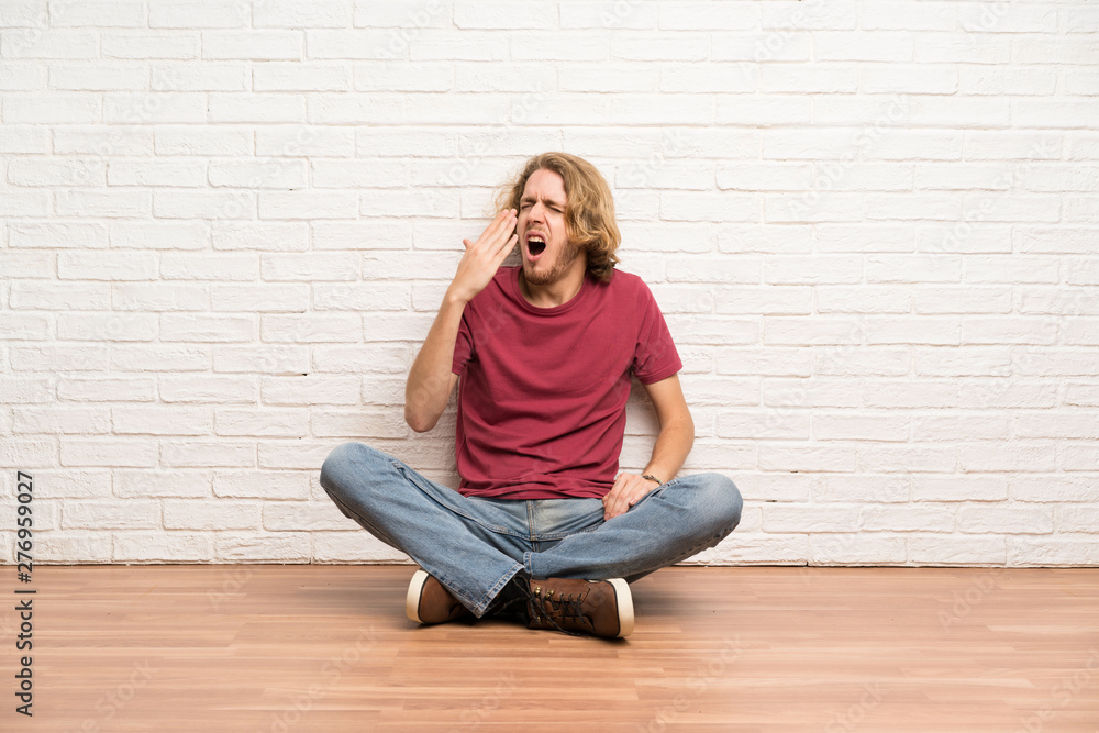 Blonde man sitting on the floor yawning and covering wide open mouth with hand Blonde man sitting on the floor yawning and covering wide open mouth with hand