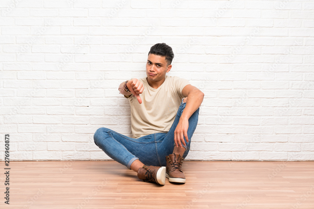 Young man sitting on the floor showing thumb down sign Young man sitting on the floor showing thumb down sign