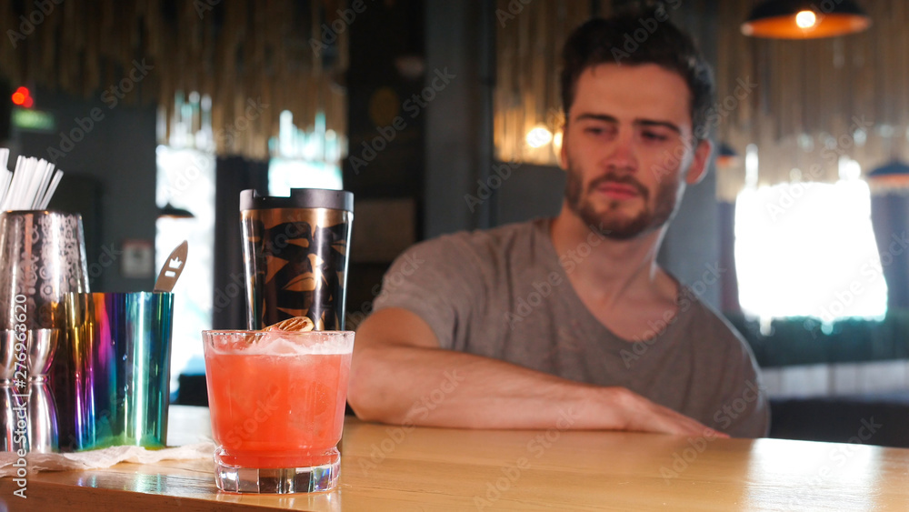 A young man sitting by the bar stand and looking at the glass with cocktail