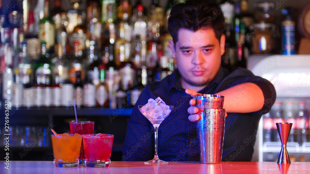 Young bartender standing in front of his bartender devices - taking a shaker - a glass with ice - cocktails on the stand