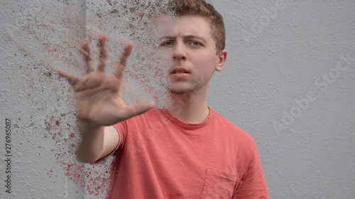 A young man wearing a bright red shirt with short hair holds out his hand desperately for help as his physical existence fades away into a million tiny particles floating away up into the sky above.