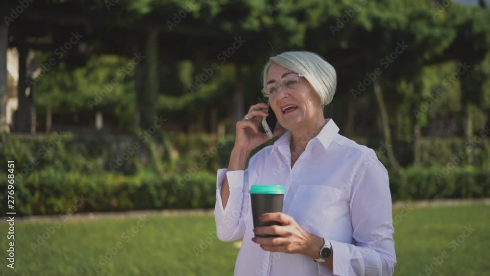 Happy senior woman talking to someone by cell phone in city park drinking coffee enjoying good conversation Happy senior woman talking to someone by cell phone in city park drinking coffee enjoying good conversation
