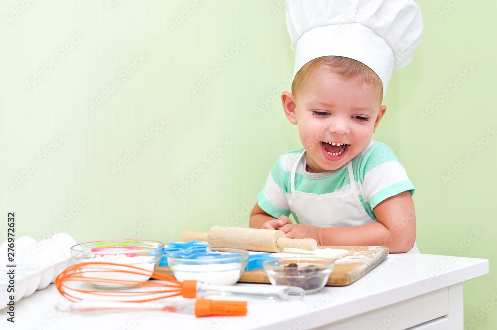 Cheerful cute baby boy in a chef costume laughs standing at the table where doing cookies. Cheerful cute baby boy in a chef costume laughs standing at the table where doing cookies.