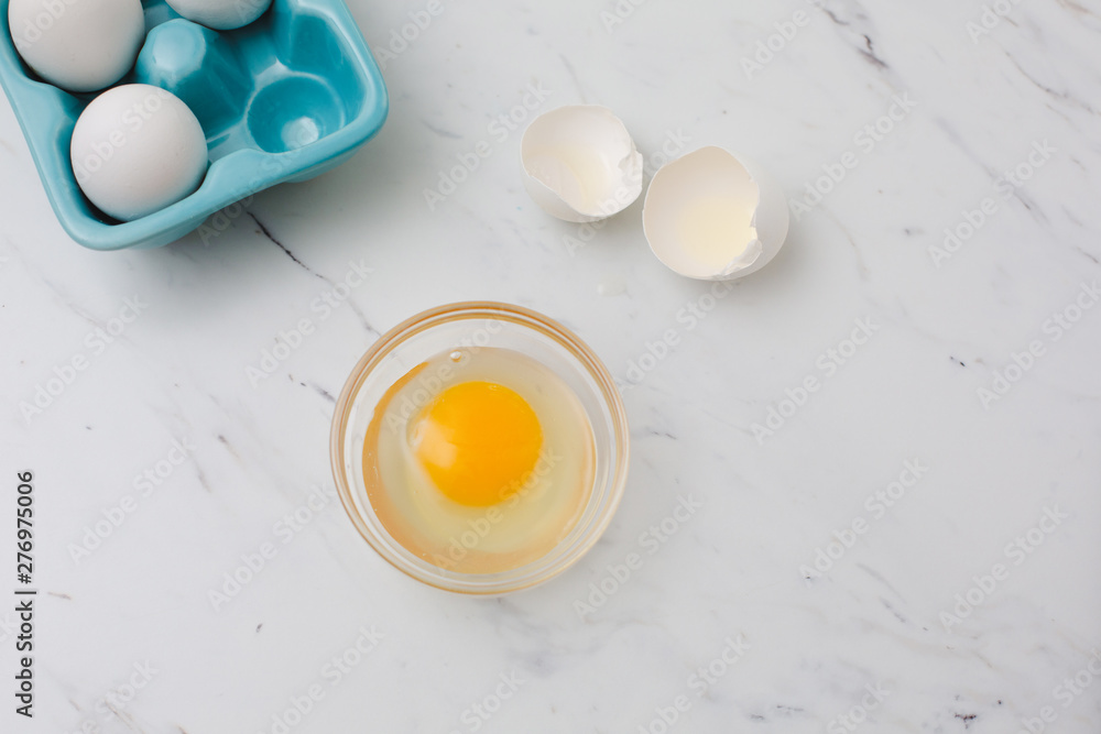 White eggs in turquoise egg tray. One egg cracked open into clear bowl on white marble countertop. White eggs in turquoise egg tray. One egg cracked open into clear bowl on white marble countertop.