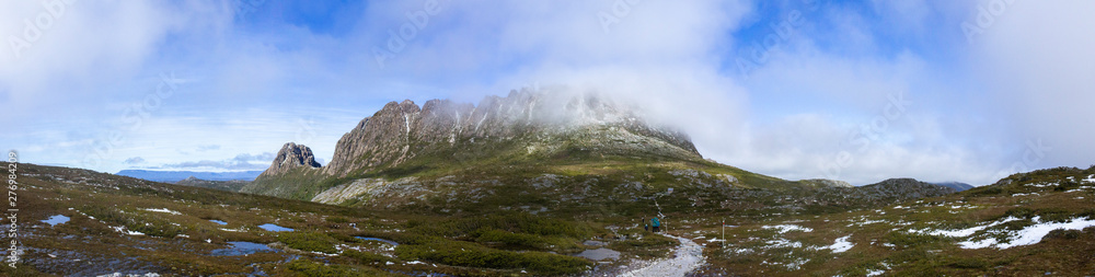 A panorama of a Cradle Mountain and Tasmania's Overland Track with fog over the summit and a hiker on the trail with some snow and water on the ground