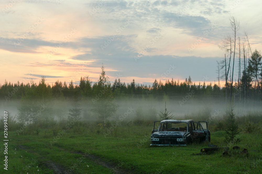 The rusty remains of the Soviet iron car lie in the forest near the road in a fog strip at sunset in the North of the Yakut taiga.