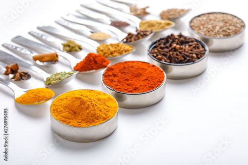 Various spices ( cumin, coriander, curry, paprika, chili, turmeric cinnamon, fenugreek, cardamom, basil leaf, parsley, cloves ) in spoons on white background. Top view with copy space.