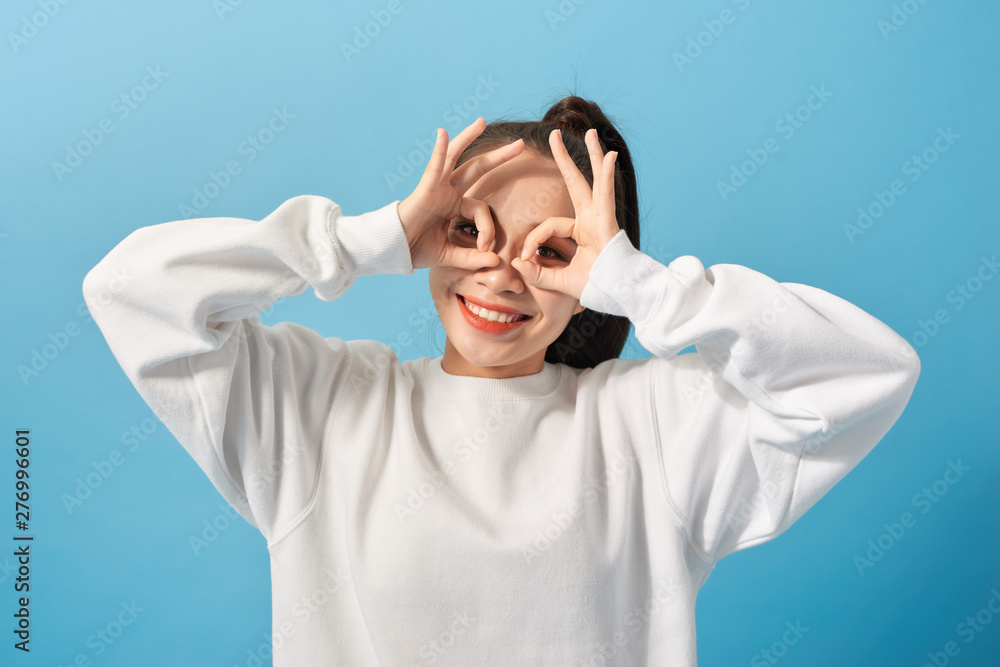 fun and people concept - smiling young woman or teenage girl looking through finger glasses over light blue background fun and people concept - smiling young woman or teenage girl looking through finger glasses over light blue background