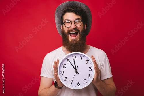 Photo of handsome excited bearded man, wearing a cowboy hat, holding clock standing over red background