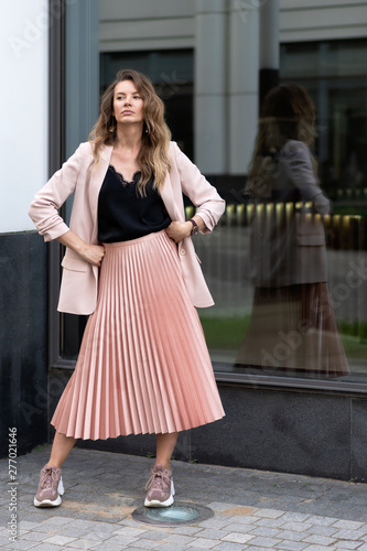 girl model in coral color clothes. Pleated long skirt, jacket, sneakers, black blouse. Long wavy hair. A very beautiful European woman stands on the street near a glass case.
