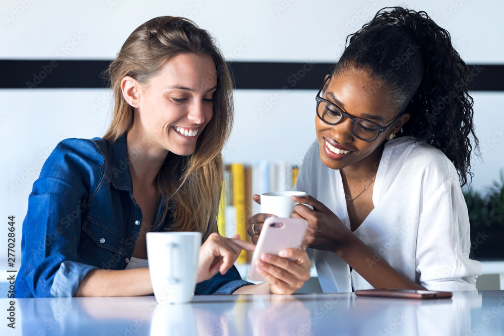 Two pretty young business women using her mobile phone together while taking a break in the office.
