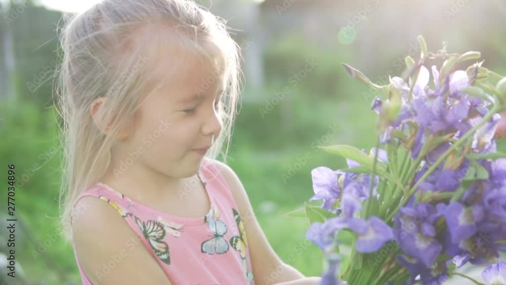 Beautiful little girl with a bouquet of blue bells stands in the garden. Baby smells blue flowers at sunset Beautiful little girl with a bouquet of blue bells stands in the garden. Baby smells blue flowers at sunset