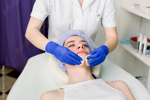 Closeup of face of woman lying on couch and relaxing during cosmetic procedure in cosmetology salon. Hands of female beautician wiping skin with cotton disks. Concept of skin care.