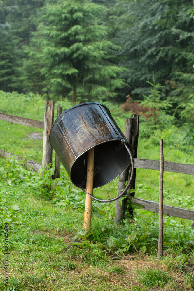 Iron bucket for the production of cheese is dried on a stick near the fence outside. The concept of natural economy, own production of cheese business in nature. Vertical Iron bucket for the production of cheese is dried on a stick near the fence outside. The concept of natural economy, own production of cheese business in nature. Vertical