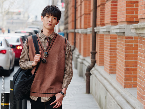 Portrait of a handsome Chinese young man with Korean style clothes walking and looking at camera with sad expression against Shanghai street background, male fashion, cool Asian young man lifestyle.