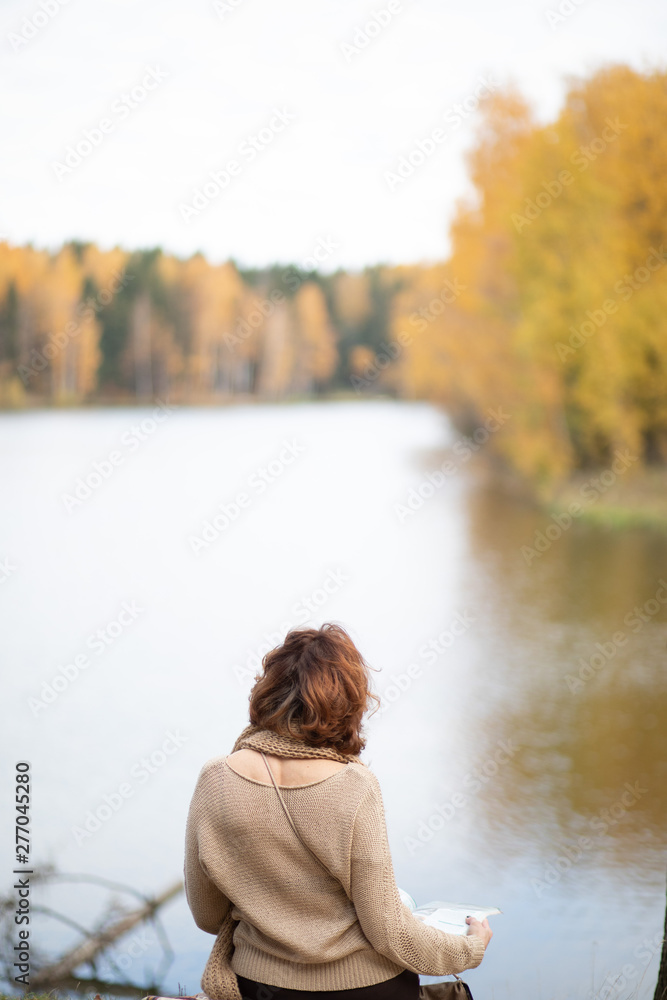 Fototapeta premium Girl sitting by the water on the lake, from the back, brunette, in beige clothes, Golden autumn.
