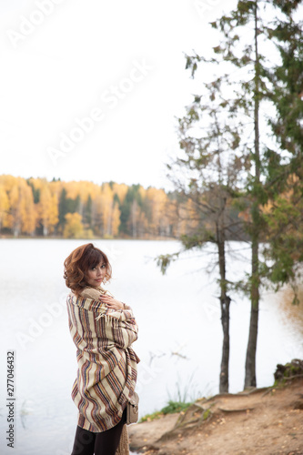 Golden autumn shooting on the lake girl stands back, brunette with curls, wrapped in a warm blanket of light brown. the scarf is knit.