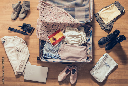 Woman's clothes, laptop, camera and flag of Spain lying on the parquet floor near and in the open suitcase. Travel concept