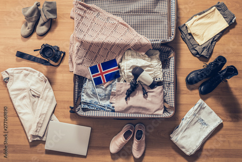 Woman's clothes, laptop, camera and flag of Iceland lying on the parquet floor near and in the open suitcase. Travel concept