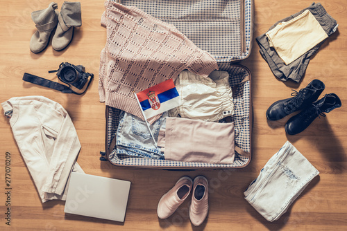 Woman's clothes, laptop, camera, russian passport and flag of Serbia lying on the parquet floor near and in the open suitcase. Travel concept