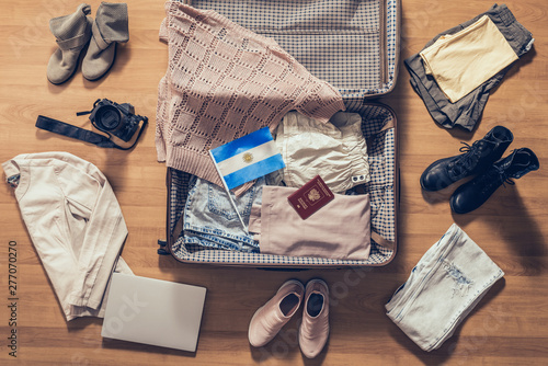 Woman's clothes, laptop, camera, russian passport and flag of Argentina lying on the parquet floor near and in the open suitcase. Travel concept