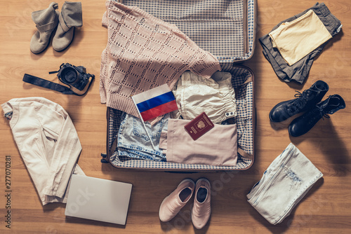 Woman's clothes, laptop, camera, russian pasport and flag lying on the parquet floor near and in the open suitcase. Travel concept