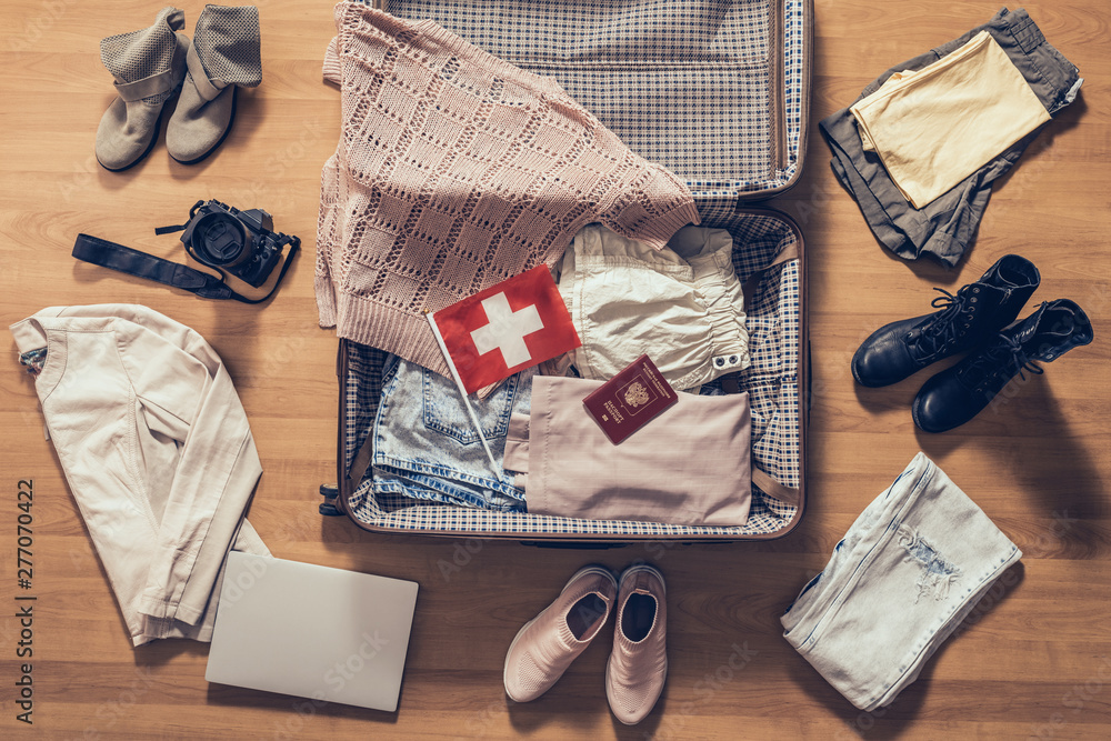 Woman's clothes, laptop, camera, russian passport and flag of Switzerland lying on the parquet floor near and in the open suitcase. Travel concept Woman's clothes, laptop, camera, russian passport and flag of Switzerland lying on the parquet floor near and in the open suitcase. Travel concept