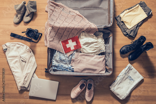 Woman's clothes, laptop, camera and flag of Switzerland lying on the parquet floor near and in the open suitcase. Travel concept