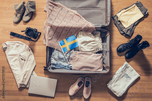 Woman's clothes, laptop, camera and flag of Sweden lying on the parquet floor near and in the open suitcase. Travel concept