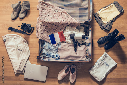 Woman's clothes, laptop, camera and flag of France lying on the parquet floor near and in the open suitcase. Travel concept
