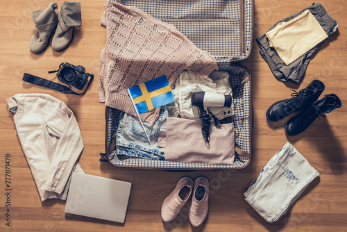 Woman's clothes, laptop, camer and flag of Sweden lying on the parquet floor near and in the open suitcase. Travel concept