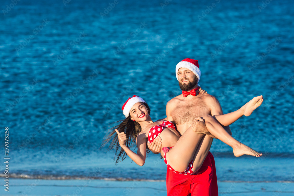 Funny bearded man and girl in Santa hats. guy holds the girl in his arms. Summer Christmas on sea beach.