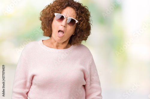 Beautiful middle ager senior woman wearing pink sweater and sunglasses over isolated background In shock face, looking skeptical and sarcastic, surprised with open mouth