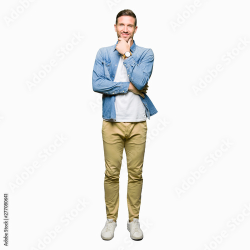 Handsome man with blue eyes and beard wearing denim jacket looking confident at the camera with smile with crossed arms and hand raised on chin. Thinking positive.