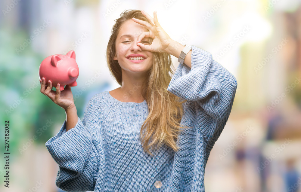 Young beautiful blonde woman holding piggy bank over isolated background with happy face smiling doing ok sign with hand on eye looking through fingers - obrazy, fototapety, plakaty 